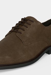 Καφέ επίσημο παπούτσι από σουέντ με σκούρα καφέ κορδόνια, ραφές και μαύρη σόλα πάνω σε λευκό φόντο.