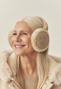 Béžové ochranné náušníky s jemnou podšívkou z umelého kožúška. Kruhový dizajn je ozdobený vyšitým obrázkom medveďa na jednej strane. Nosené osobou s dlhými vlasmi.