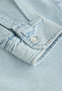 Manche de veste en denim bleu clair avec fermeture à boutons blancs et coutures visibles sur les plis du tissu.