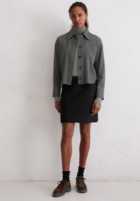 Grijze, korte button-up jasje over een lichtgrijze coltrui, gecombineerd met een zwarte rok. Een gestructureerde stof en bruine veterschoenen maken de look compleet.