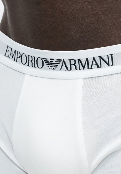 Κοντινό πλάνο στη λευκή ζώνη της εσωρούχης Emporio Armani σε σκούρο δέρμα, με μαύρο λογότυπο και κείμενο στη ελαστική ταινία.