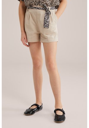 Kind trägt beige Shorts mit Leopardenmuster-Krawattengürtel, Leopardenmuster-Top und schwarze Lack-Mary-Jane-Schuhe, steht mit den Händen in den Taschen.