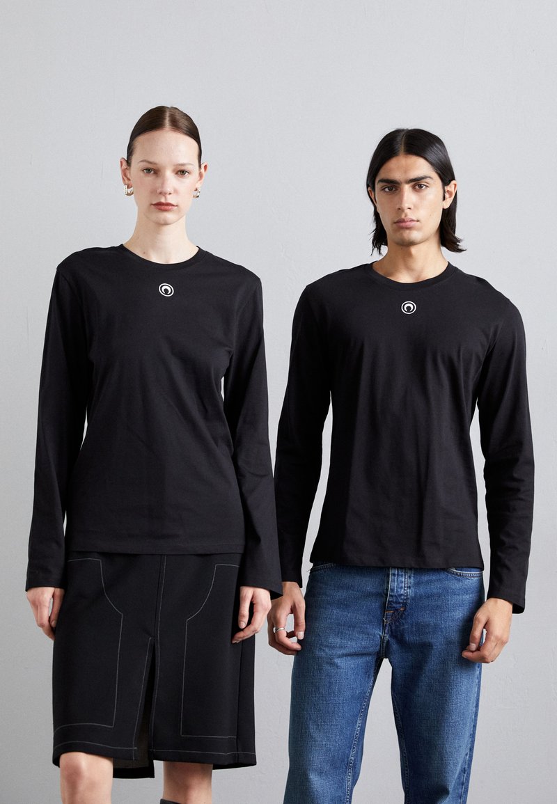 Črne dolge majice z dolgimi rokavi in okroglo logo na prsih, v kombinaciji s črno krilom in modrimi jeans hlačami, v minimalističnem okolju.