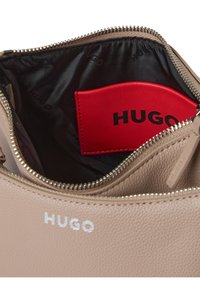 Beige Handtasche aus Leder mit einem Reißverschluss, ausgestattet mit einem schwarzen Innenfutter und einem roten HUGO-Tag. Silberne Markenkennzeichnung an der Außenseite.
