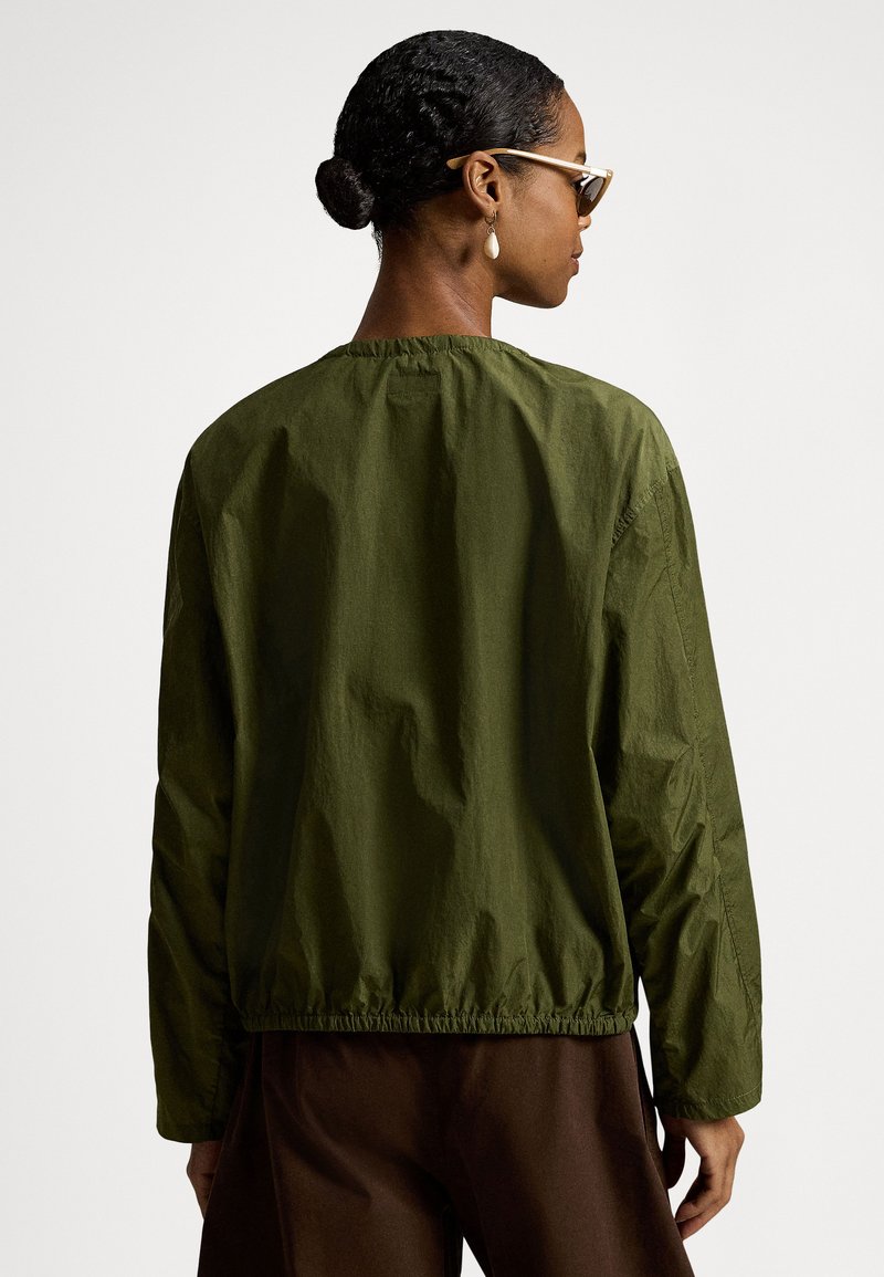 Woman with dark hair in bun wearing tan sunglasses, teardrop earrings, olive green long-sleeve top, and brown pants, facing away against white background.