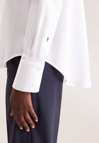 White long-sleeve shirt with a curved hem, button cuffs, and embroidered floral detail on cuff, paired with navy pants.