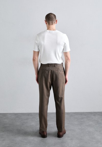 Chemise blanche à manches courtes et pantalons texturés marron avec poches arrière, dotés d'une coupe décontractée et de bas de pantalon retroussés. Des chaussures marron complètent le look.