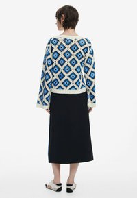 Pull en crochet avec un motif en diamant bleu et crème, associé à une longue jupe noire et des chaussures claires. Tissu tricoté texturé.