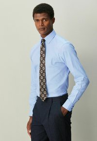 Lyseblå skjorte med lange ærmer, kombineret med et mørkebrunt slips med geometrisk mønster. Navyblå bukser med nålestriber fuldender outfittet.