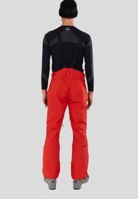 Červené lyžařské kalhoty s nastavitelnými popruhy, voděodolnou látkou, širokým střihem nohavic a zesílenými manžetami. Kombinovány s černým dlouhým rukávem a šedými lyžařskými botami.