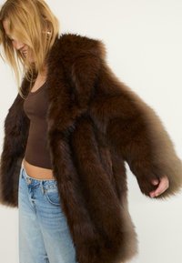 Cappotto in pelliccia sintetica marrone con ampio colletto e maniche oversize, indossato sopra un top marrone, abbinato a jeans azzurri chiari. Materiale piacevole al tatto e peloso.