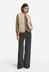 Beige vadderad väst med kabelstickad mönster, bärs över en brun tröja, ihop med svarta vida byxor och taupe ankle boots.