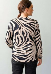 Frau mit braunen Haaren, die zu einem Pferdeschwanz gebunden sind, trägt eine schwarze und beige Bluse mit Zebramuster und schwarze Hosen und steht vor einer schlichten weißen Wand.