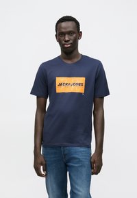 Μπλε βαμβακερό t-shirt με ορθογώνιο πορτοκαλί γραφικό που αναγράφει "JACK & JONES" με μαύρα γράμματα, λαιμόκοψη και κοντά μανίκια.