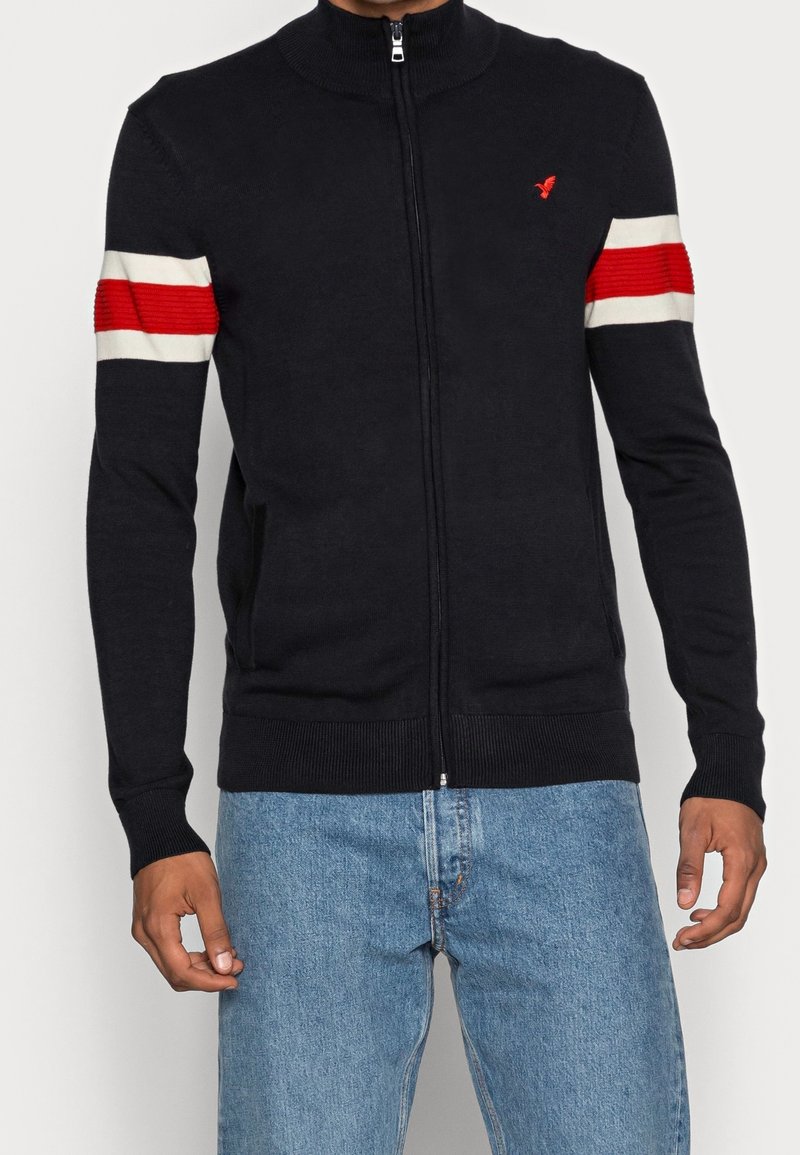 Marinblå zip-up cardigan med röda och vita randiga detaljer på ärmarna, ribbade muddar och krage. Tillverkad av ett mjukt, texturerat stickat tyg.