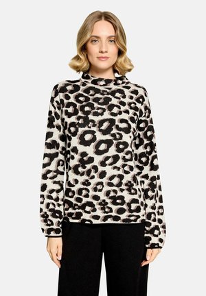 Kvinde med kort blondt hår iført en langærmet leopardmønstret top og sorte bukser, stående mod en ensfarvet lys baggrund.
