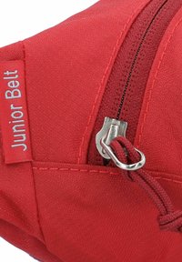 Rode stoffen zak met een rits, voorzien van een label met de tekst "Junior Belt." Het materiaal heeft een gestructureerd oppervlak.