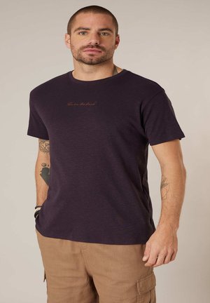 Man in een donkerpaarse T-shirt met kleine tekst en beige cargobroek tegen een effen neutrale achtergrond.