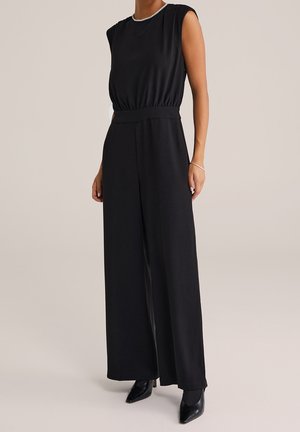 Combinaison noire sans manches avec taille froncée et jambes larges, portée avec des talons noirs à bout pointu et un bracelet argenté au poignet droit.
