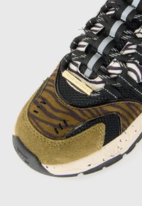 Gros plan sur l’avant d’une basket avec un tissu rayé tigre, du daim olive, un mesh noir, des lacets noirs et blancs, et un détail de plaque dorée avec le nom.