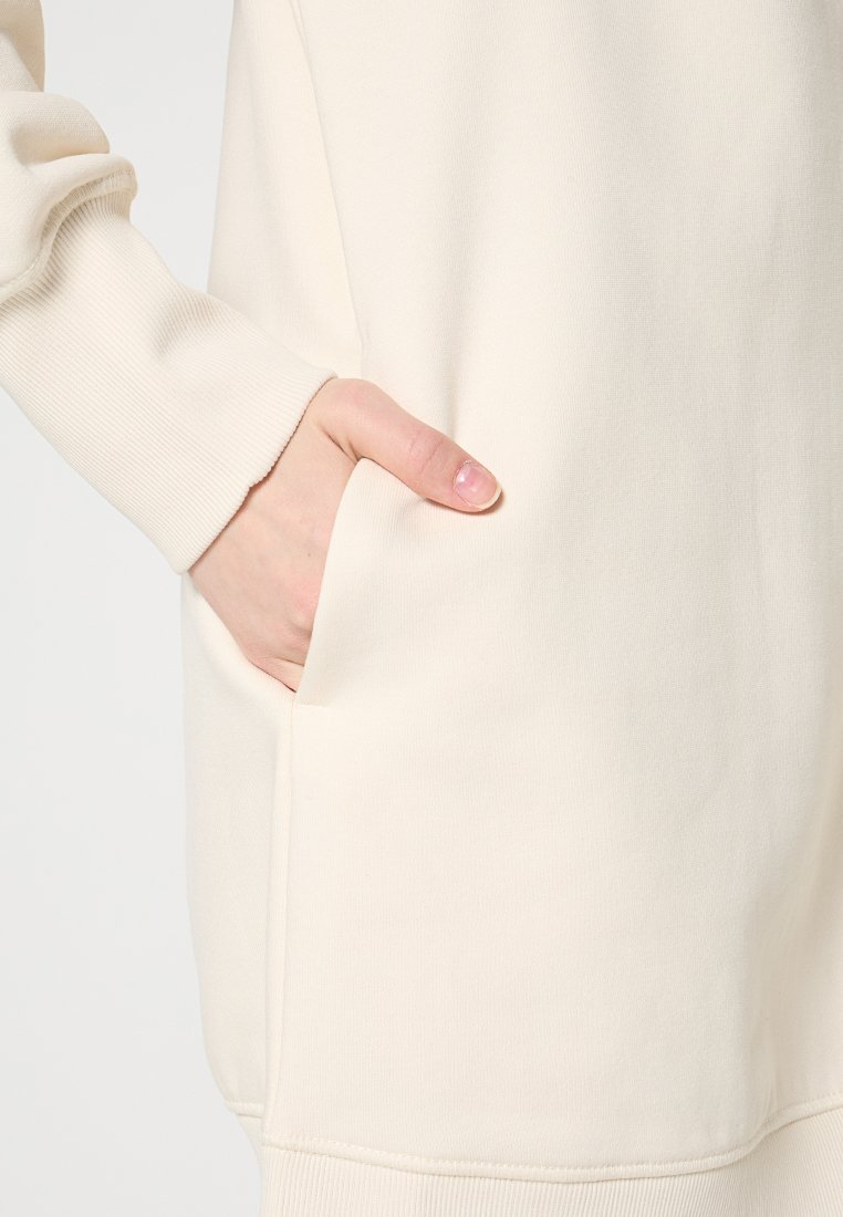 Cremefarbener Pullover mit strukturiertem Stoff und sichtbarer Fronttasche. Abgerundeter Saum und lockere Passform.