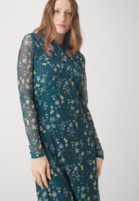 Pitkähihainen mekko tummassa teal-värissä kukkakuviolla, jossa on teksturoitu päällikerros ja laskostus yksityiskohtana kaula-aukossa. Pehmeä, puoliksi läpinäkyvä kangas.
