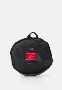 Svart mesh duffelbag med rund form, med et rødt merke som viser "BASE CAMP DUFFEL L" og glidelås. Håndtak på toppen.