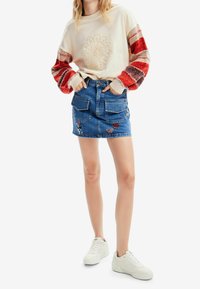 Kremowy sweter z teksturowym, haftowanym wzorem i czerwonymi paskami na rękawach, zestawiony z niebieską spódnicą dżinsową z haftem floralnym i kieszeniami.