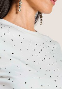 Weißes Oberteil mit glatter Textur, verziert mit kleinen schwarzen Edelsteinen in verschiedenen Größen. Verfügt über einen runden Ausschnitt und minimale Designelemente.