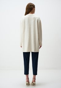 Cardigan lungo a maniche, di colore crema, lavorato a maglia con una finitura morbida e testurizzata, indossato sopra pantaloni tapered blu navy. La vista posteriore mostra una vestibilità rilassata e orlo a costine.