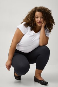 Witte korte mouwen t-shirt, marineblauwe legging en zwarte flats. Krullend bruin haar, knielende pose met hand op de kin. Eenvoudige achtergrond.