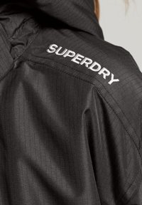 Zwarte jas met een gestructureerd patroon, voorzien van een capuchon en een geborduurd "SUPERDRY" logo op de rechter schouder in het wit.