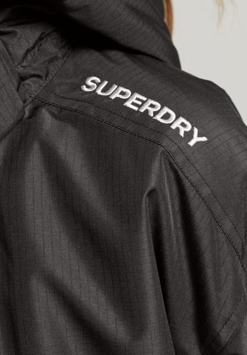 Zwarte jas met een gestructureerd patroon, voorzien van een capuchon en een geborduurd "SUPERDRY" logo op de rechter schouder in het wit.