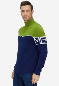 Homme portant un pull demi-zip vert et marine avec des lettres blanches sur le haut du bras, associé à un jean bleu, posant devant un fond blanc.