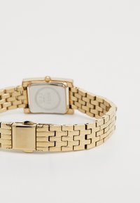 Montre en métal doré avec un boîtier carré, un bracelet à maillons et une fermeture. Dos gravé avec le logo de la marque et les détails de certification.