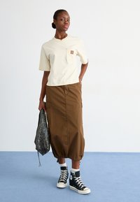 Beige katoenen t-shirt met een borstzak, gecombineerd met een bruine rok tot halverwege de kuiten met cargozakken en een geplooide zoom. Zwarte high-top sneakers.