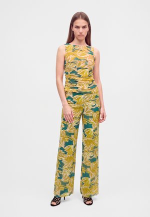 Femme debout portant un haut sans manches et un pantalon large avec de grands motifs floraux jaunes et verts ainsi que des talons ouverts noirs.
