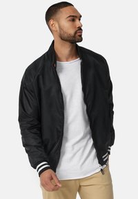 Chaqueta bomber negra en tela suave y lisa. Cuenta con un cierre frontal, puños de rayas blancas y negras acanalados, y un bolsillo con cremallera en el costado.