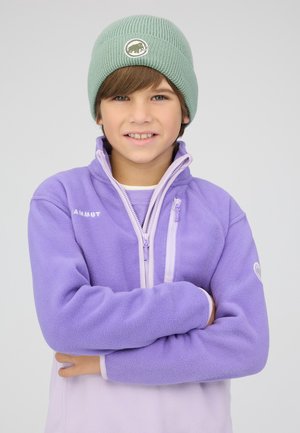 Ragazzo con le braccia incrociate che indossa un berretto verde lavorato a maglia e una giacca in pile viola Mammut su uno sfondo grigio uniforme.