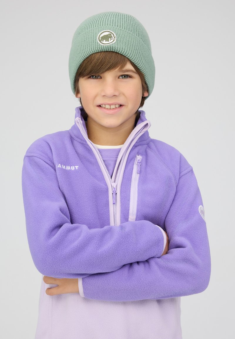 Garçon avec les bras croisés portant un bonnet en tricot vert et une veste polaire Mammut violette sur un fond gris uni.