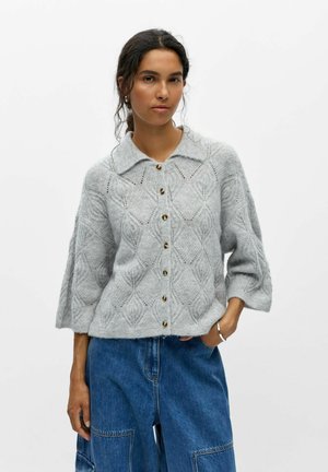 Femme portant un cardigan gris en maille boutonné avec des motifs en losange texturés et des manches larges, associé à un pantalon en denim bleu.