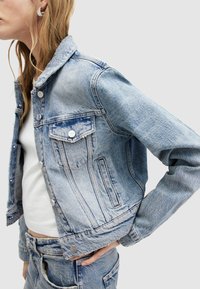 Giacca di jeans azzurro chiaro con un design aderente, dotata di bottoni argentati, tasche laterali e un colletto classico. Dettaglio del tessuto testurizzato visibile.