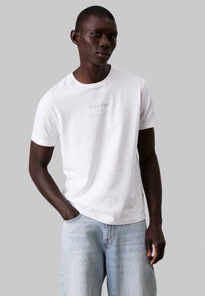 Hvid bomulds-T-shirt med rund halsudskæring, prydet med et lysegrå Calvin Klein-logo på brystet. Båret sammen med lyseblå denimjeans.