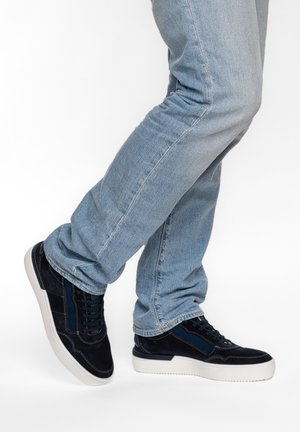 Person trägt hellblaue Jeans und schwarze Sneakers mit weißen Sohlen, steht mit einem Fuß leicht angehoben vor einem einfarbigen weißen Hintergrund.