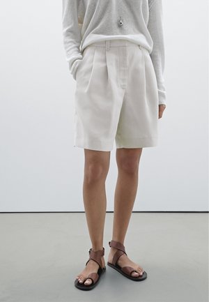Personne portant un short plissé crème, un pull léger, un collier pendentif et des sandales en cuir marron avec bride à la cheville, debout sur un sol gris.