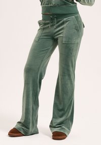 Samtgrüne Hose mit lockerer Passform, ausgestattet mit Seitentaschen und einem Kordelzugbund. Kombiniert mit braunen, weichen Hausschuhen.