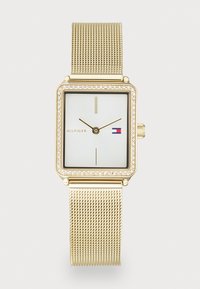 Montre bracelet carrée dorée avec bracelet en maille, lunette en cristal, cadran blanc, marqueurs d'heure minimalistes, et logo Tommy Hilfiger sur le visage.