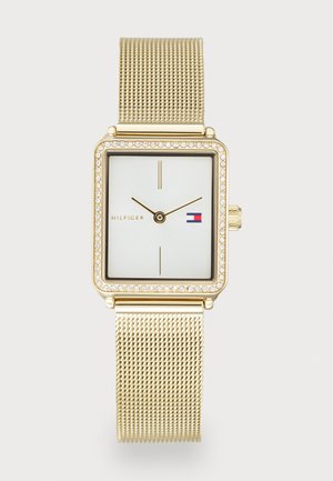 Montre bracelet carrée dorée avec bracelet en maille, lunette en cristal, cadran blanc, marqueurs d'heure minimalistes, et logo Tommy Hilfiger sur le visage.