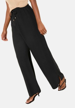 Vrouw draagt zwarte wijde broek met trekkoord in de taille en zijsplitten, beige mouwloze top en zwarte sandalen met hak.