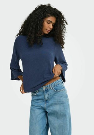 Jeune femme aux cheveux bouclés portant un haut bleu foncé ample et un jean taille haute bleu clair, soulevant légèrement son haut.